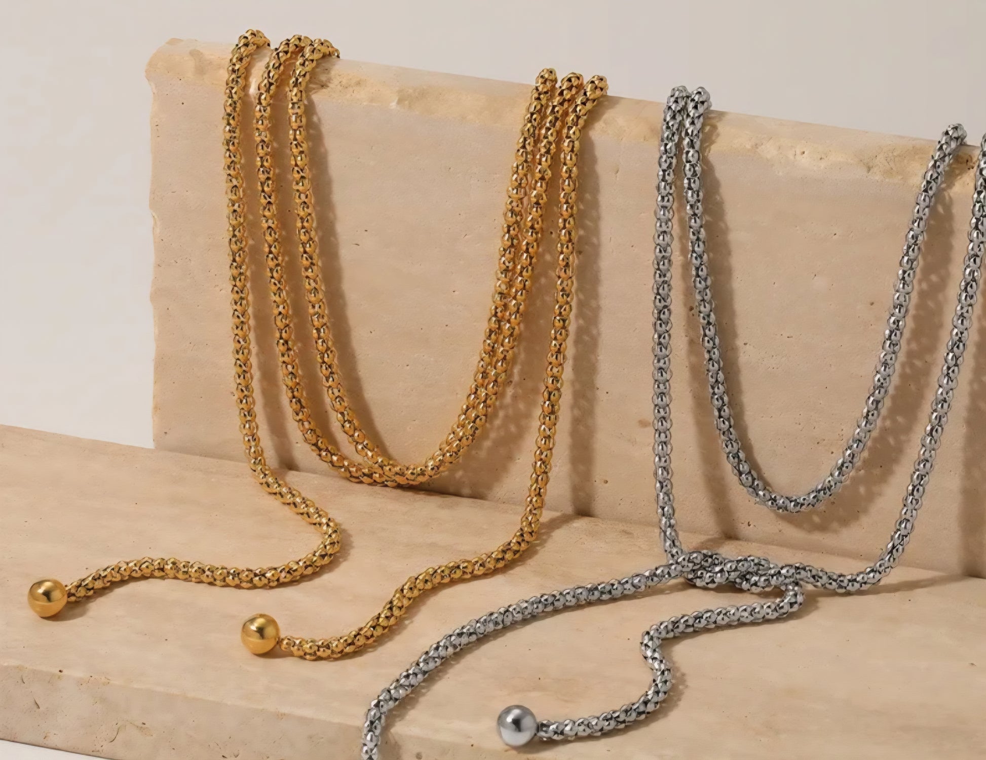 One long gold chain and one long silver chain draped over a beige surface.