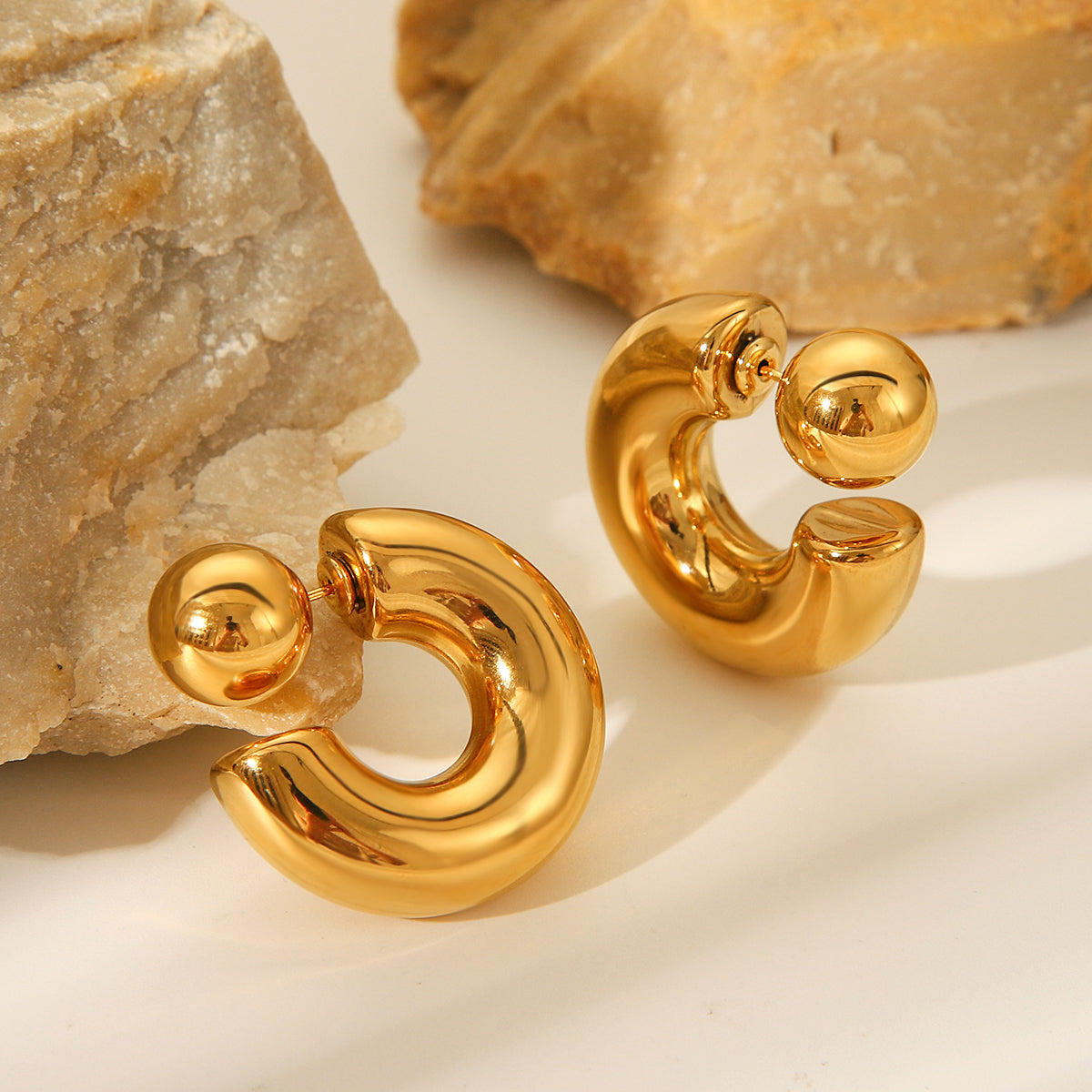 Gold hoop earrings on a textured beige surface with rocks in the background