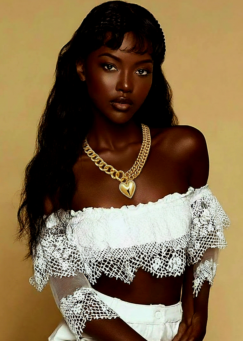 Woman wearing a white lace top and gold necklace with heart-shaped pendant against a beige background.