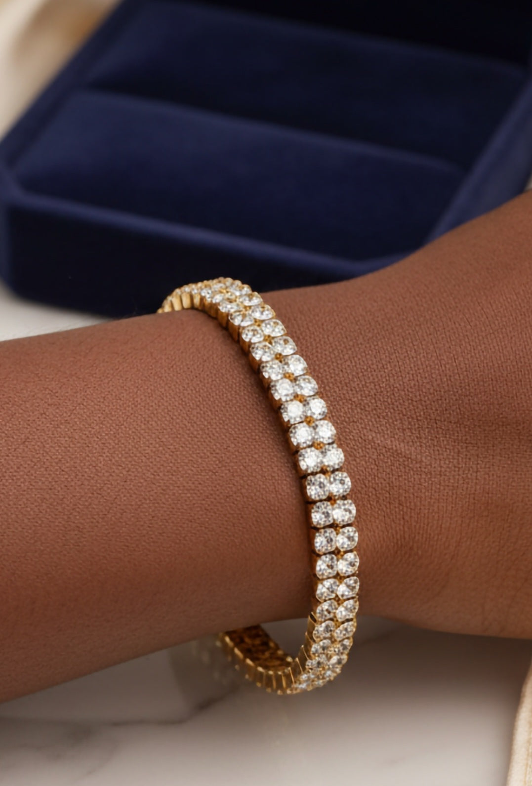 Gold Tennis bracelet on a wrist against a dark blue jewelry box background