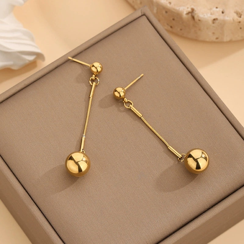 Gold dangling earrings with a gold ball at the end on a beige jewelry box with a neutral background