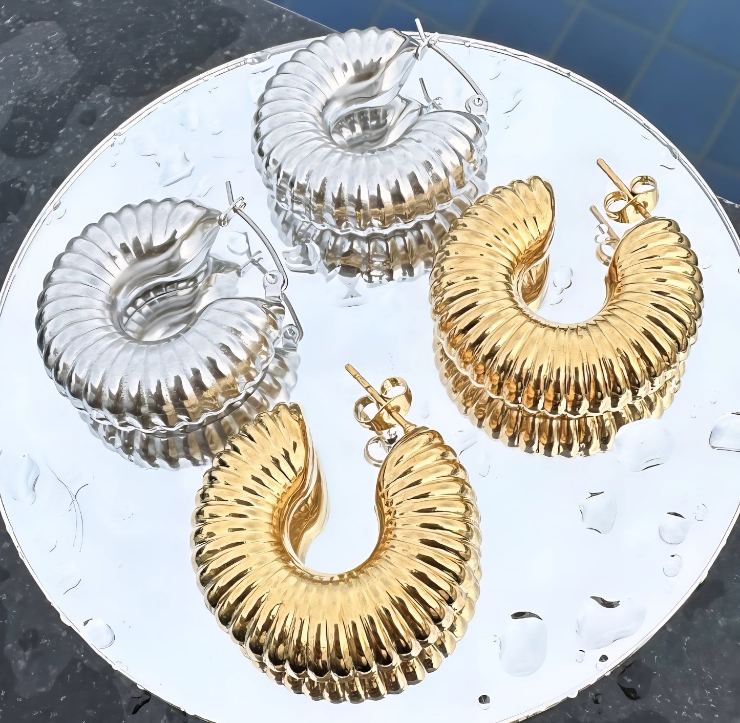 Gold and silver hoop earrings on a round metallic surface with water droplets.