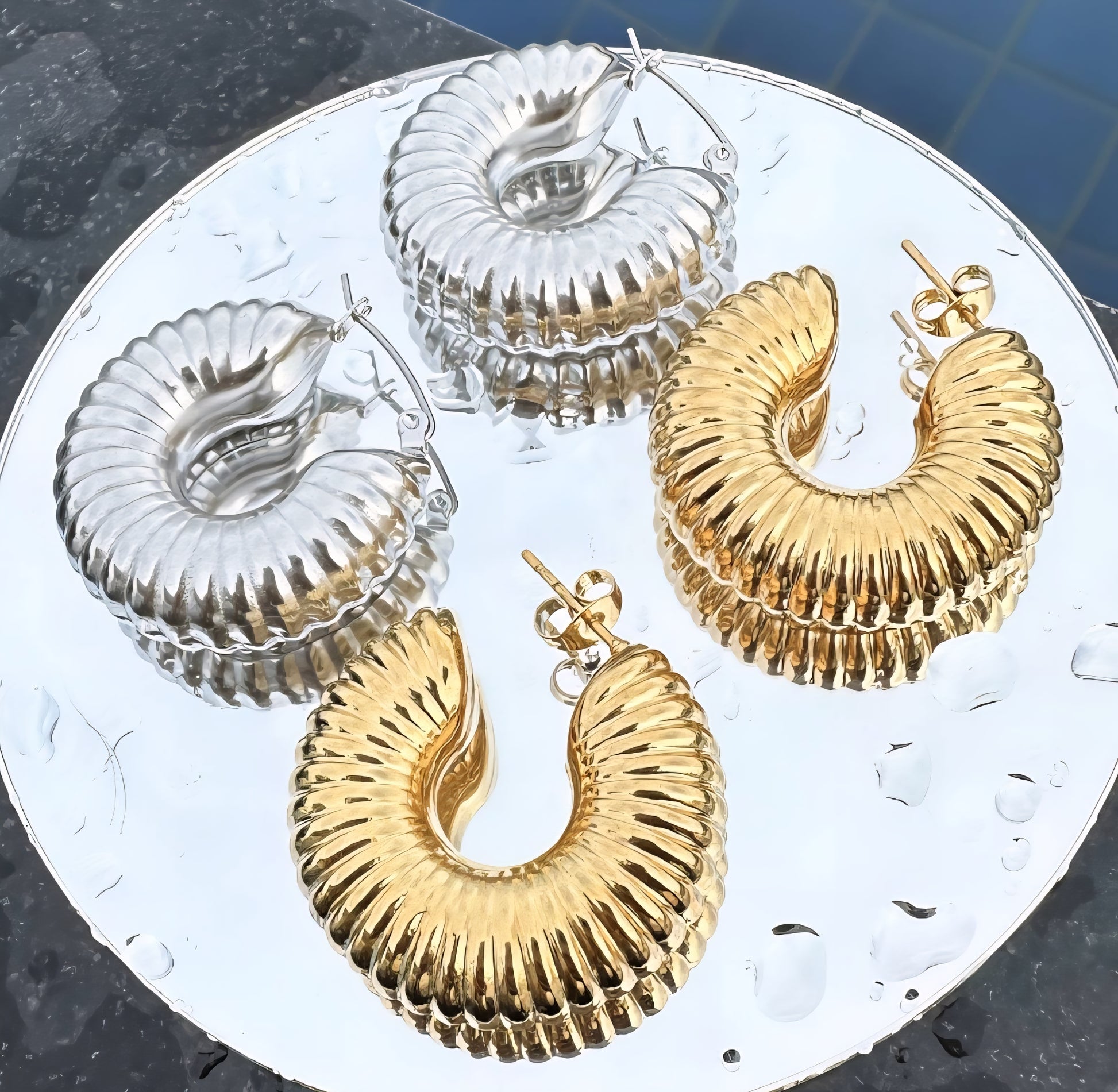 Gold and silver hoop earrings on a round metallic surface with water droplets.